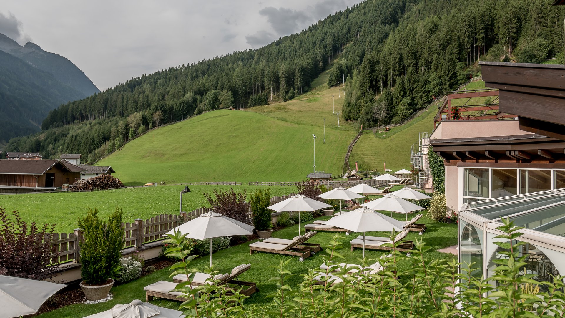 Stubaital: das Hotel mit Pool Liegestühle mit Sonnenschirmen im Garten vor Bergwald und grünen Hügeln