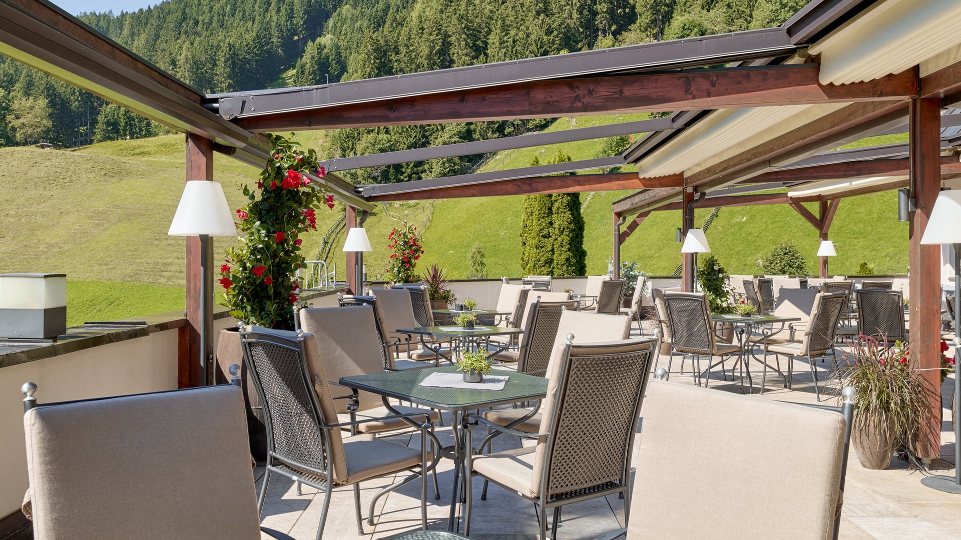 Stubaital: das Hotel mit Pool Terrasse mit Stühlen und Tischen vor bewaldetem Hügel im Sonnenschein