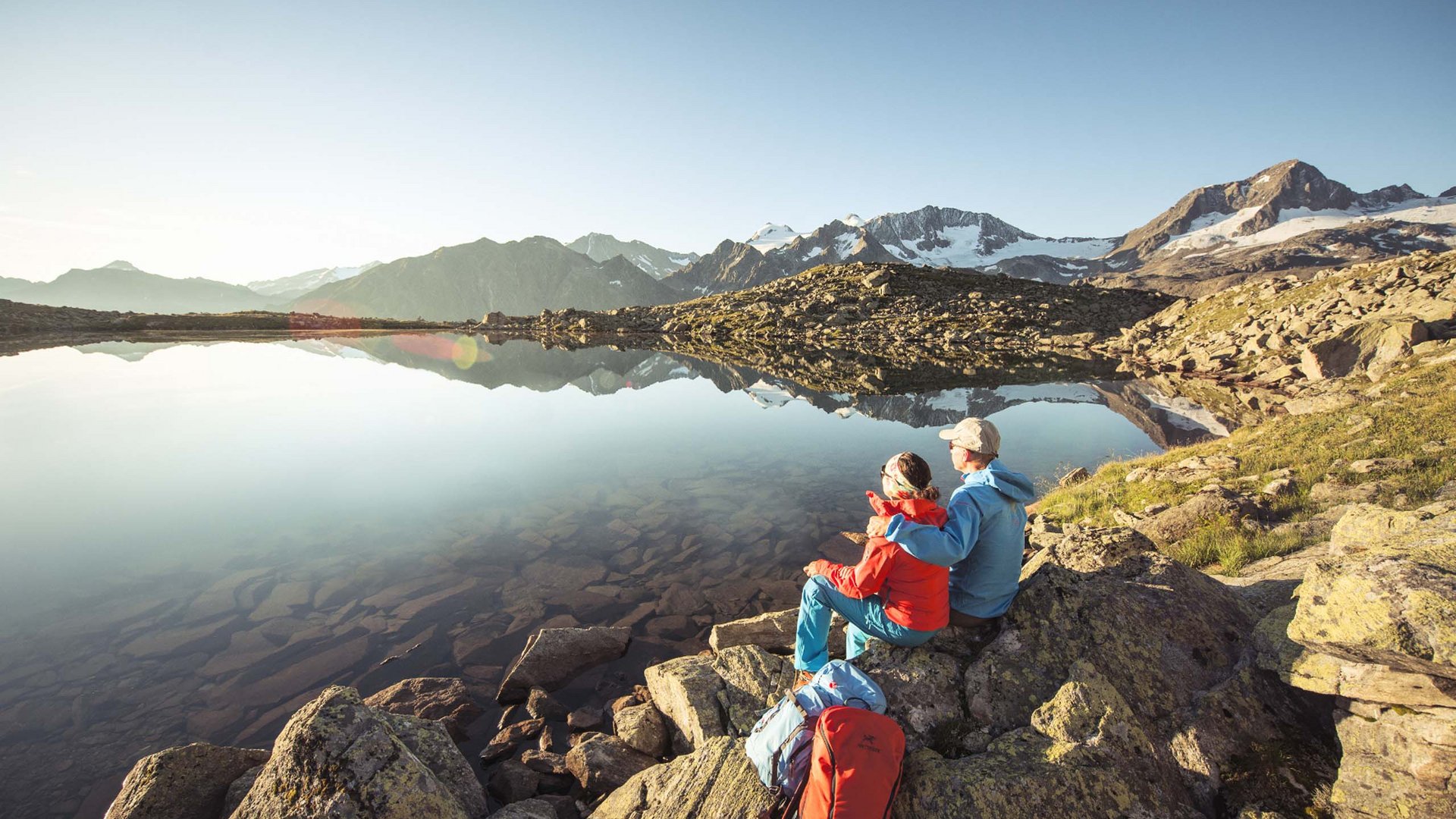 Stubaital: das Hotel mit Pool Zwei Wanderer sitzen auf Felsen und blicken auf einen ruhigen Bergsee bei Sonnenaufgang